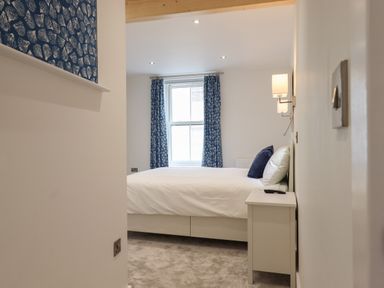 A bedroom with a bed white bedding blue pillows bedside table window with blue curtains and wall lamps at Ferryside in Kingswear
