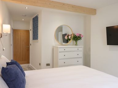 A bedroom with a wooden door white dresser a round mirror pink flowers and a wall-mounted TV at Ferryside in Kingswear