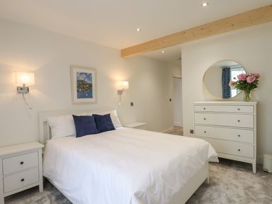 A bedroom with a double bed with white bedding and blue pillows beside two white nightstands and a white dresser with a round mirror and flowers at Ferryside Kingswear