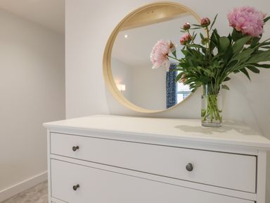 A white dresser with drawers a round mirror above and a vase of pink flowers on top at Ferryside in Kingswear