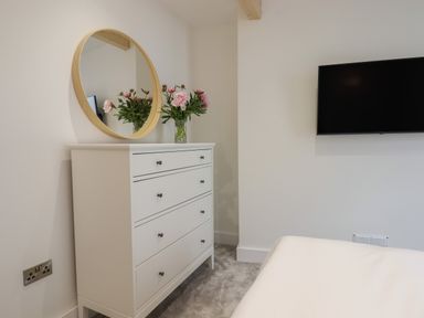 A bedroom with a white dresser with a round mirror and flowers on top and a wall mounted television at Ferryside in Kingswear