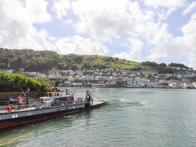 A ferry carrying vehicles across a river with houses and trees on a hillside in the background at Ferryside in Kingswear