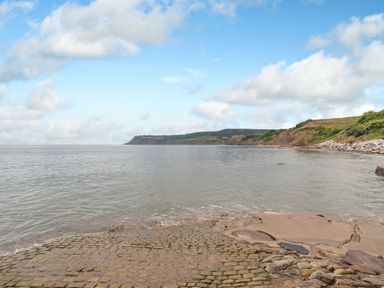 A beach with cobblestone and concrete shore and a cliff in the distance at The Shepherds Rest in Robin Hoods Bay