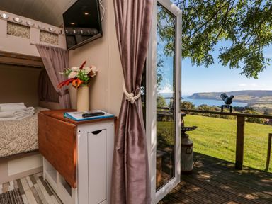 A small bedroom with bunk beds flowers on a cabinet and a wall-mounted tv opening onto a deck with a view of a grassy field and coastline at The Shepherd's Rest in Robin Hood's Bay