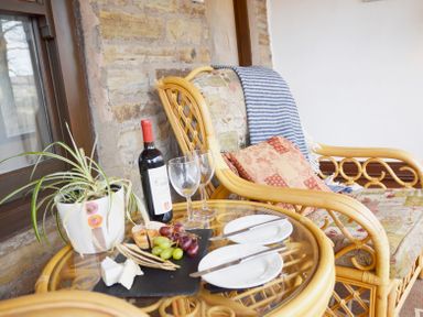 A wicker armchair with cushions and a striped blanket next to a glass table with wine, glasses, cheese, grapes, and a plant at Keepers in Nenthead near Alston