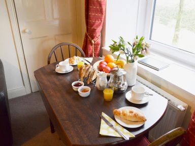 A dining table set with croissants cups of tea or coffee glasses of orange juice jam bowls a fruit bowl flowers in a vase by a window at Keepers Nenthead near Alston