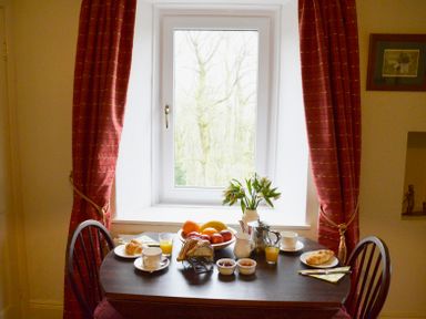 A small dining table set with fruit, croissants, jam, juice, cups, and a flower vase in front of a window with red curtains at Keepers in Nenthead near Alston