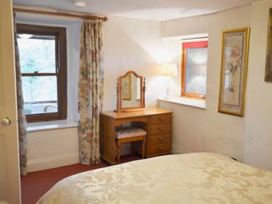 A bedroom with a wooden dressing table and stool next to two windows with floral curtains at Keepers in Nenthead near Alston