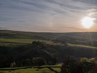 A landscape with green fields trees and hills under a sky with the sun at Keepers Nenthead near Alston