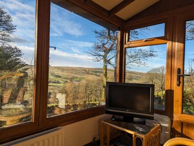 A small room with large windows showing a countryside view and a television on a wicker table at Keepers in Nenthead near Alston