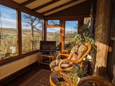 A sunroom with large windows overlooking a landscape with a chair a small table with plants and a television at Keepers in Nenthead near Alston