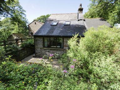 A stone cottage with a slate roof surrounded by green plants and flowers at Bothy in Nenthall near Alston