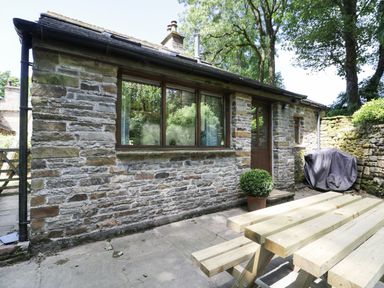 The exterior of a stone building with a wooden door and window a potted plant a covered barbecue and a wooden picnic table on a stone patio at Bothy in Nenthall near Alston