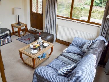 A living room with a leather sofa covered with blue striped throws a glass top coffee table with snacks a lamp on a wicker side table and a large window at Bothy in Nenthall near Alston