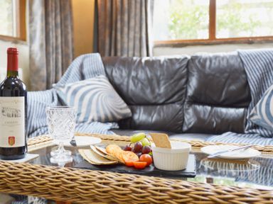 A glass coffee table with a bottle of red wine crackers fruits and a dip in front of a black leather sofa with pillows and curtains at Bothy in Nenthall near Alston