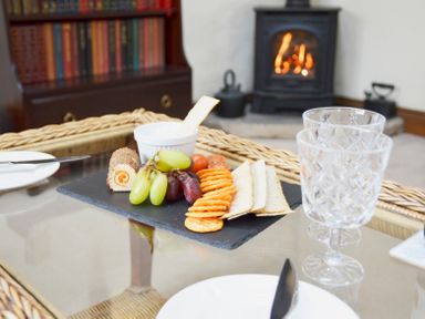A dining table with snacks on a slate board and two crystal glasses in a room with a fireplace and a bookshelf at Bothy in Nenthall near Alston