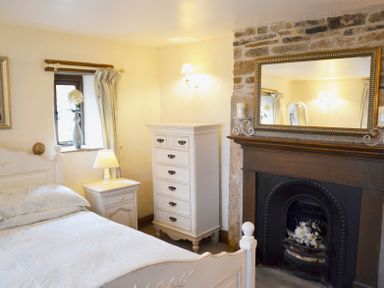 A bedroom with a bed a bedside table with lamp a chest of drawers and a fireplace with a mirror above at Bothy in Nenthall near Alston