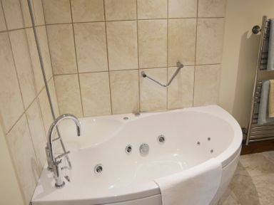 A bathroom with a white bathtub with jets a chrome faucet a hand shower a towel on the edge tiled walls and a towel rack with towels at Bothy in Nenthall near Alston