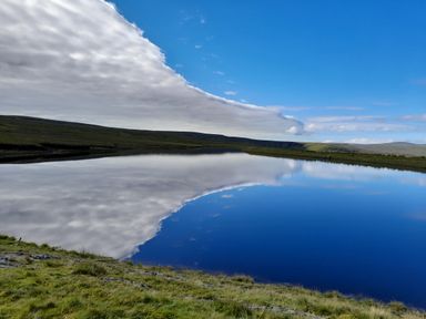 A lake reflecting clouds and blue sky surrounded by grassy hills at Bothy in Nenthall near Alston