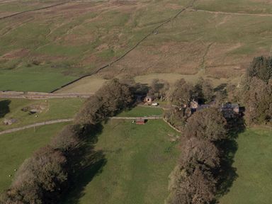 An aerial view of a rural farmstead with several buildings surrounded by fields and trees at Bothy in Nenthall near Alston