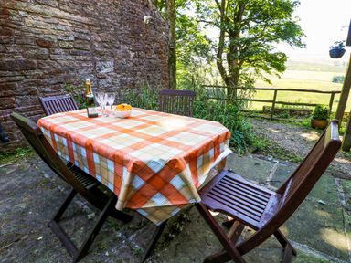 An outdoor patio with a table covered by an orange checkered tablecloth surrounded by four wooden chairs with a bottle and glasses on the table at Chestnut in Nenthall near Alston