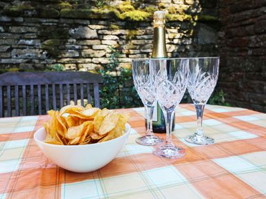 A bowl of potato chips with a bottle of champagne and three crystal glasses on a table outdoors with a stone wall background at Chestnut in Nenthall near Alston