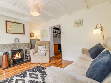 A living room with a beige armchair and sofa with cushions a lit fireplace a lamp and a patterned rug at Vi's Valley View in Pentewan