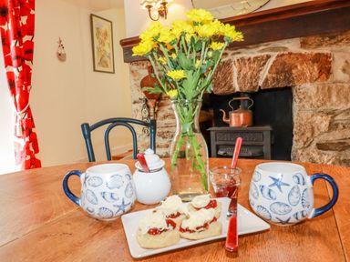 A dining table with mugs with shell patterns scones with cream and jam a vase with yellow flowers and a stone fireplace in the background at Vi's Valley View in Pentewan