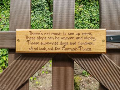 A wooden gate with a carved wooden sign warning about uneven steps and Cornish Pixies surrounded by green leaves
