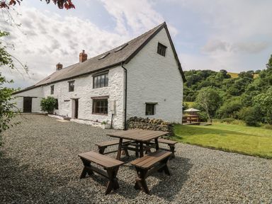 An outdoor seating area with wooden picnic benches and a white stone house with windows and chimneys at Penarth in Cregrina near Builth Wells