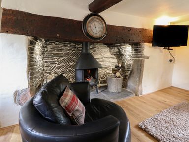 A living room with a round black leather chair a wood burning stove stone wall a clock above the fireplace a basket with logs a mounted television and a light colored rug at Penarth in Cregrina near Builth Wells
