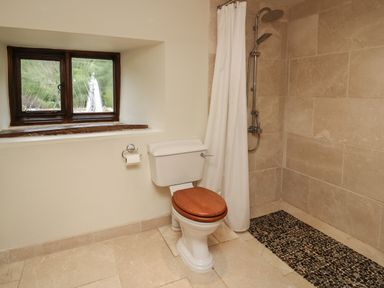 A bathroom with a toilet with a wooden seat and a walk-in shower with a curtain and pebble floor at Penarth in Cregrina near Builth Wells