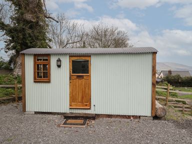 A small corrugated metal hut with a wooden door and window on a gravel surface at Rabbit Hill Hut in Govilon