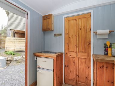 A small kitchen area with a fridge, wooden cabinets, and a door at Rabbit Hill Hut in Govilon