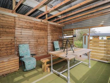 An outdoor covered seating area with two cushioned chairs a wooden table with a telescope on it a bench and wooden panel walls at Rabbit Hill Hut in Govilon