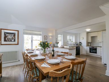 A dining room with a wooden table set for a meal and an open kitchen in the background at The Old School House in Lower Swell near Stow-On-The-Wold