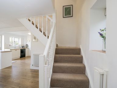 A staircase with carpeted steps and white railing next to a kitchen with wooden floor and white cabinets at The Old School House in Lower Swell near Stow-On-The-Wold