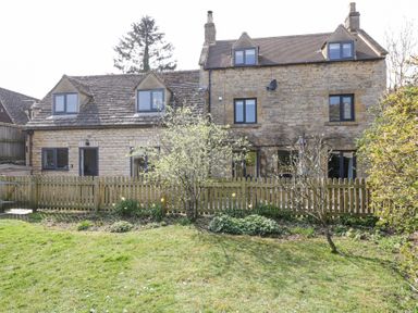 A stone house with dormer windows a wooden fence and a garden with trees and grass at The Old School House in Lower Swell near Stow-On-The-Wold