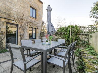 An outdoor patio area with a table and eight chairs a closed umbrella champagne a vase with flowers at The Old School House in Lower Swell near Stow-On-The-Wold