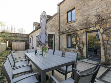 An outdoor patio with a table and chairs a closed umbrella flowers and a bottle of champagne at The Old School House in Lower Swell near Stow-On-The-Wold
