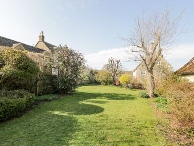 A garden with grass, trees, bushes, stone buildings, and a table with two chairs at The Old School House in Lower Swell near Stow-On-The-Wold