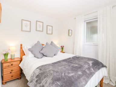 A bedroom with a double bed with gray pillows and blanket two wooden nightstands with lamps and flowers a window with white curtains at Thornhills Cottage in Othery near Middlezoy