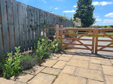 A wooden gate with a gray wooden fence and flowers along the fence next to a paved area at Thornhills Cottage in Othery near Middlezoy