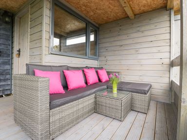 An outdoor seating area with a wicker sofa with gray cushions and pink pillows and a small glass table with flowers at Thornhills Lodge Othery near Middlezoy