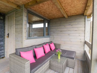A covered porch with a wicker sectional sofa with pink cushions and a small glass table with flowers at Thornhills Lodge in Othery near Middlezoy