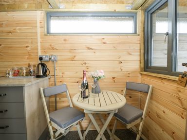 A small dining area with two cushioned chairs a round table with a bottle and wine glasses beside a countertop with a kettle and jars at Thornhills Lodge in Othery near Middlezoy