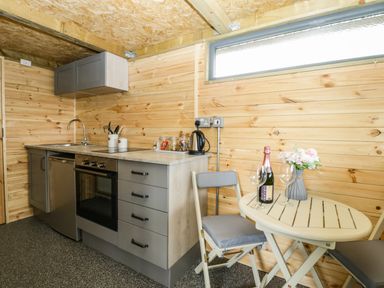 A small kitchen with wooden walls a stove a sink a kettle jars utensils a round table with two chairs flowers and a bottle of champagne at Thornhills Lodge in Othery near Middlezoy