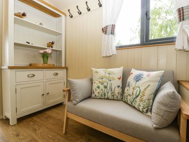 A small sitting area with a light grey sofa and floral cushions beside a white cabinet with shelves and a window with white curtains at Thornhills Lodge in Othery near Middlezoy