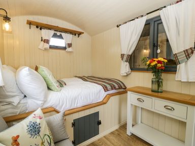 A small bedroom with a bed, pillows, a window with curtains, a white table with drawers, and a vase of flowers at Thornhills Lodge in Othery near Middlezoy