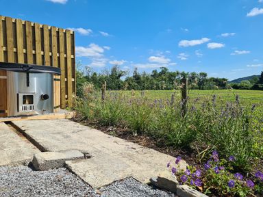 A garden with a wooden hot tub on a paved area next to a flower bed and a green field at Thornhills Lodge in Othery near Middlezoy